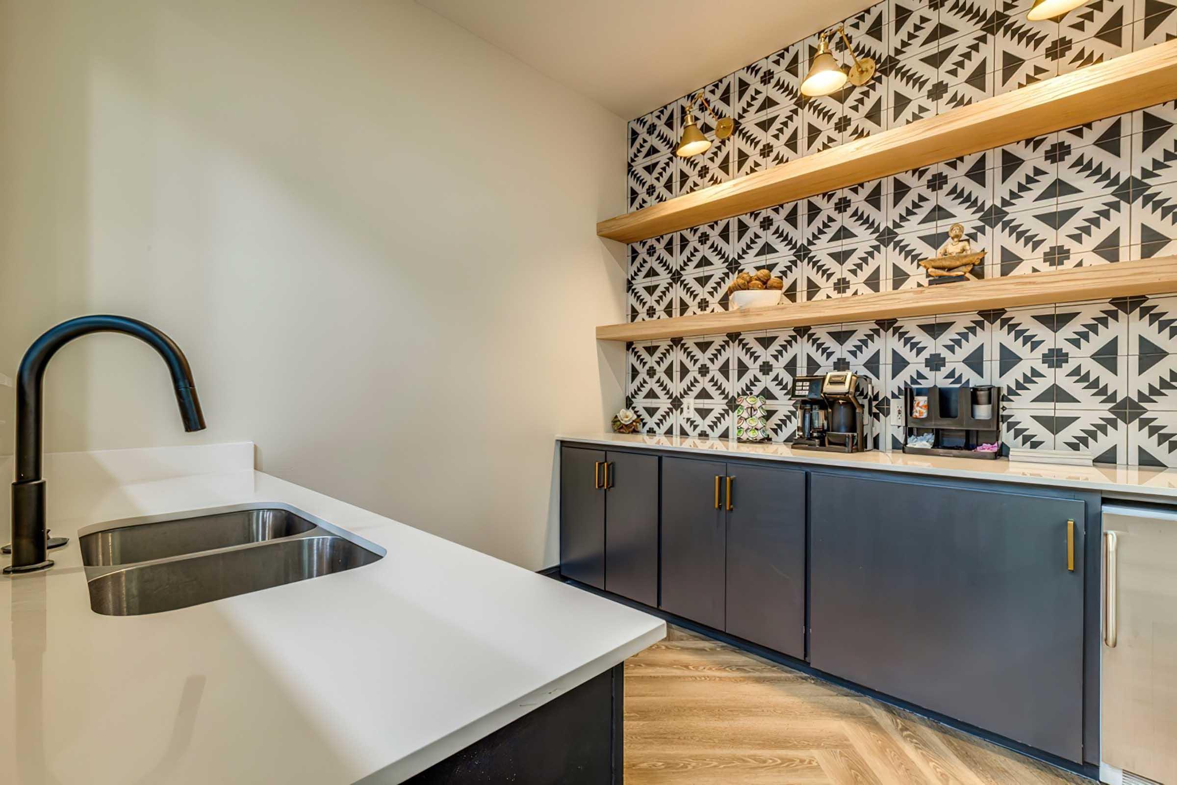 A modern kitchen featuring a sleek black sink and countertop. The wall behind is decorated with a geometric patterned wallpaper, and there are two wooden shelves displaying decorative items. Below, a dark cabinet holds a coffee maker, suggesting a stylish and functional space.