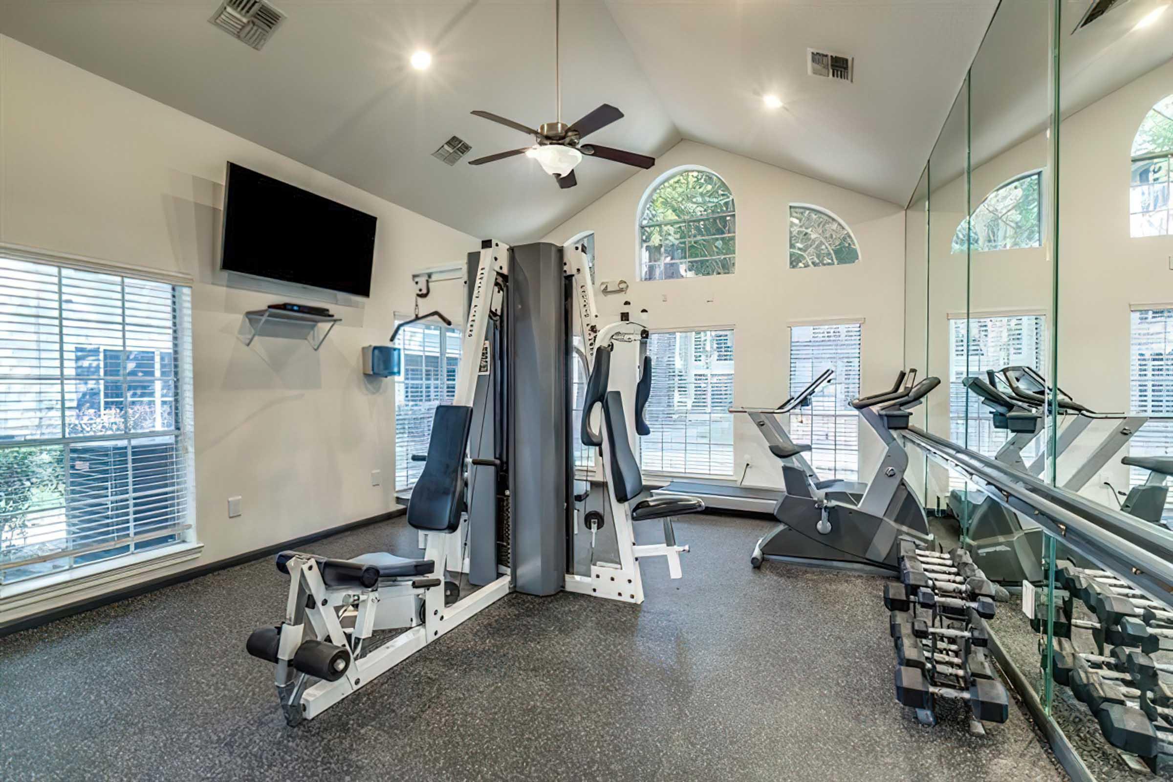 A modern gym featuring a multi-station weight machine, a treadmill, and a set of free weights. The room has large windows allowing natural light in, a ceiling fan, and mirrors on one wall, creating an open and inviting workout space.