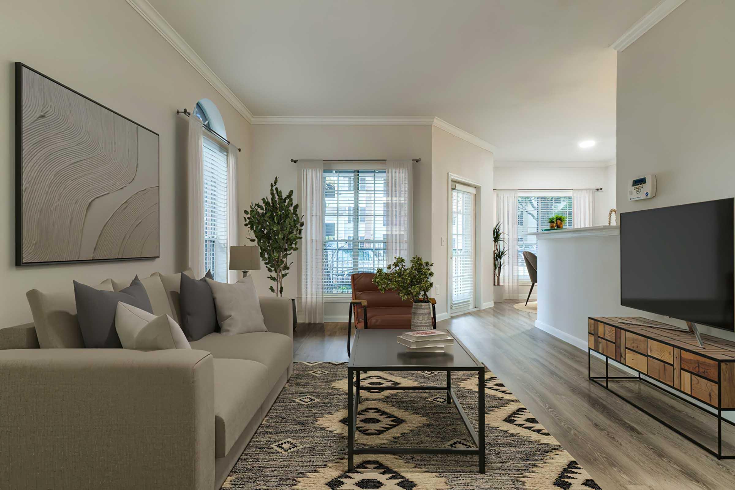 A modern living room featuring a light gray sofa adorned with decorative pillows, a black coffee table, and an area rug with geometric patterns. Natural light flows through large windows with white blinds. A plant stands in the corner, and a side chair complements the space. A kitchen area is visible in the background.