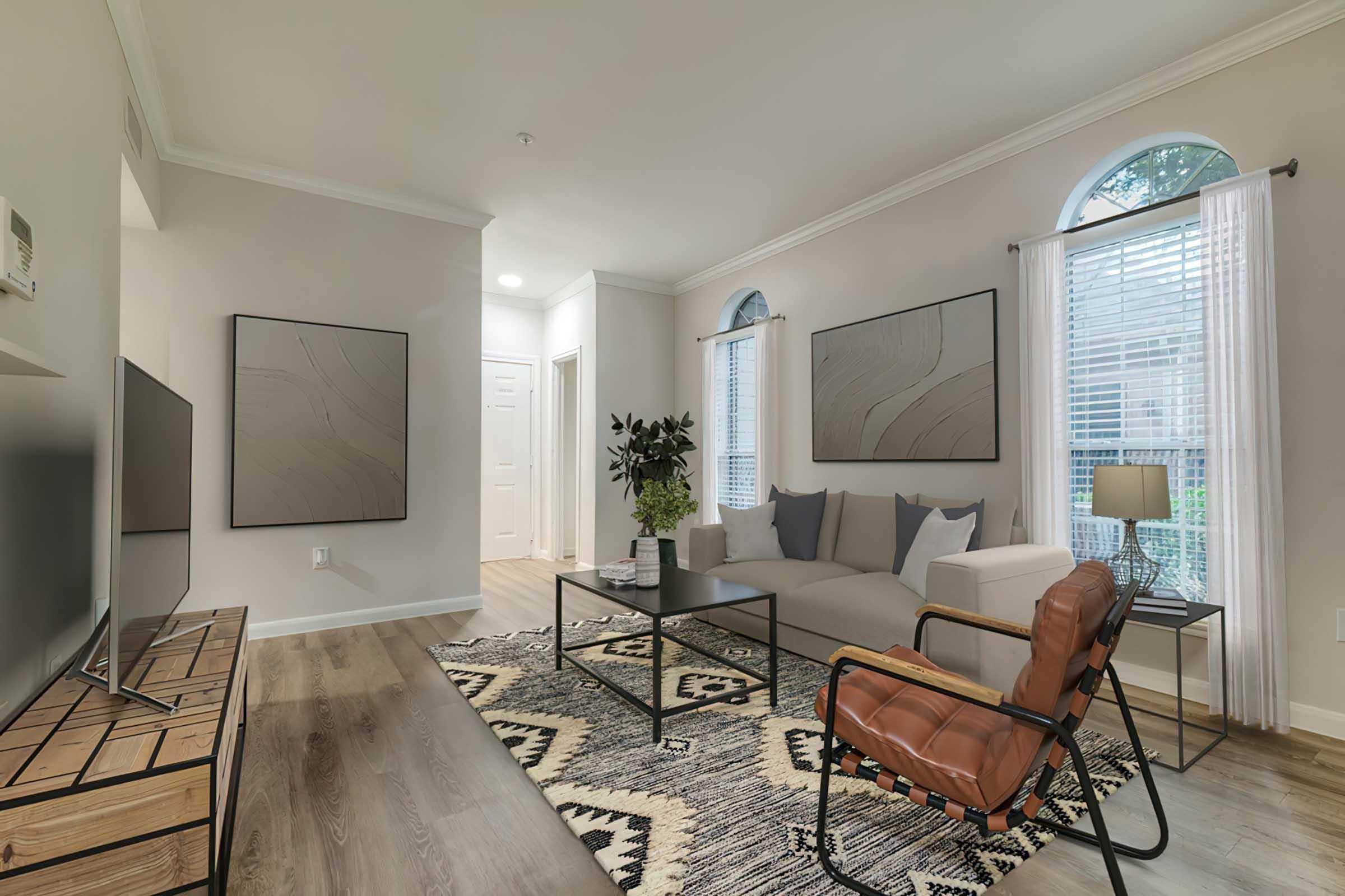A modern living room with neutral-colored walls, featuring a light gray sofa, a brown leather chair, a black coffee table, and a patterned area rug. Two framed artworks hang on the walls, and large windows allow natural light to brighten the space. A potted plant adds a touch of greenery.