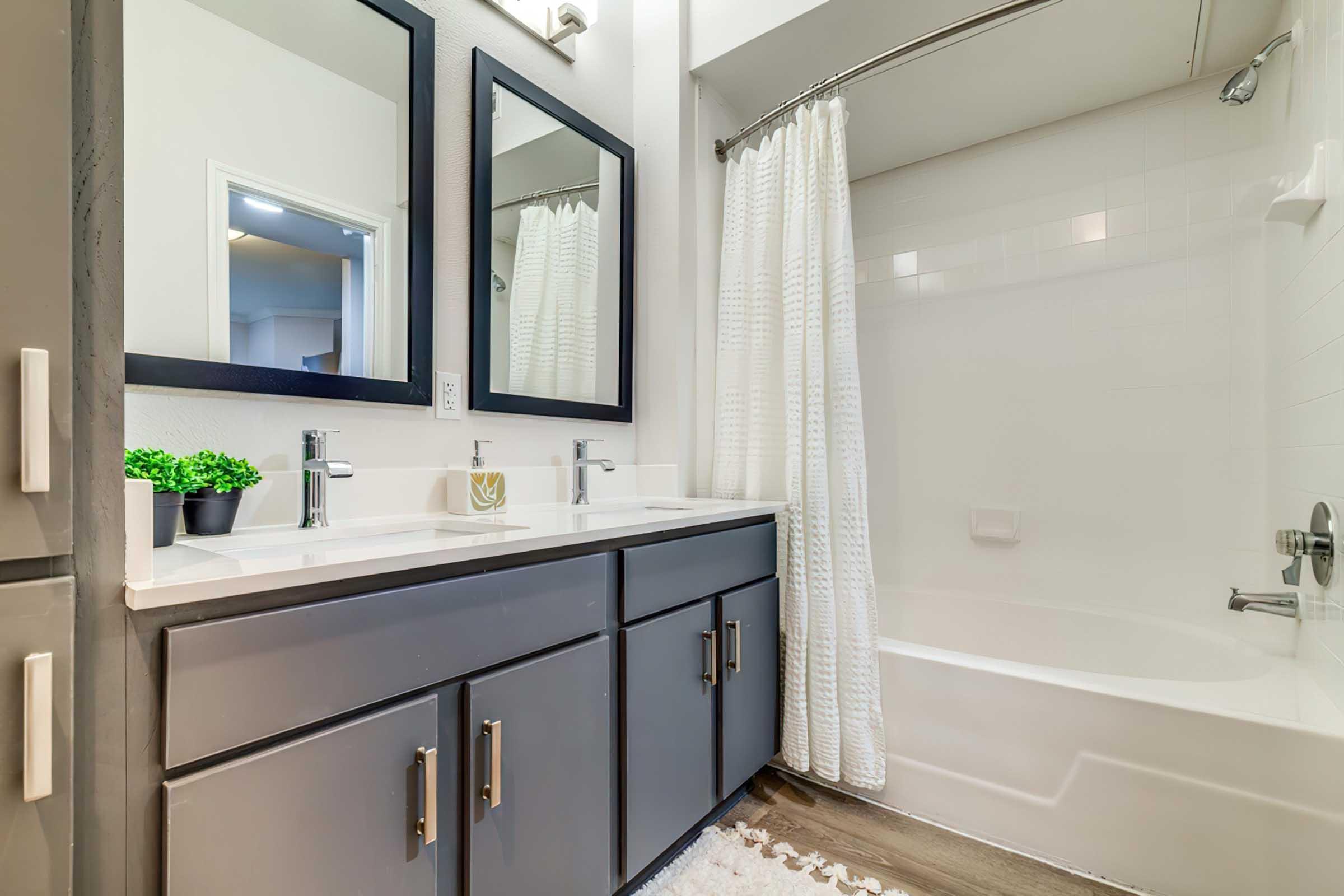 A modern bathroom featuring a double sink vanity with dark cabinetry, two sleek mirrors, and a white tiled shower area with a curtain. A small potted plant adds a touch of greenery, and the floor is covered with a soft rug. Natural light is seen in the adjacent room through a door.