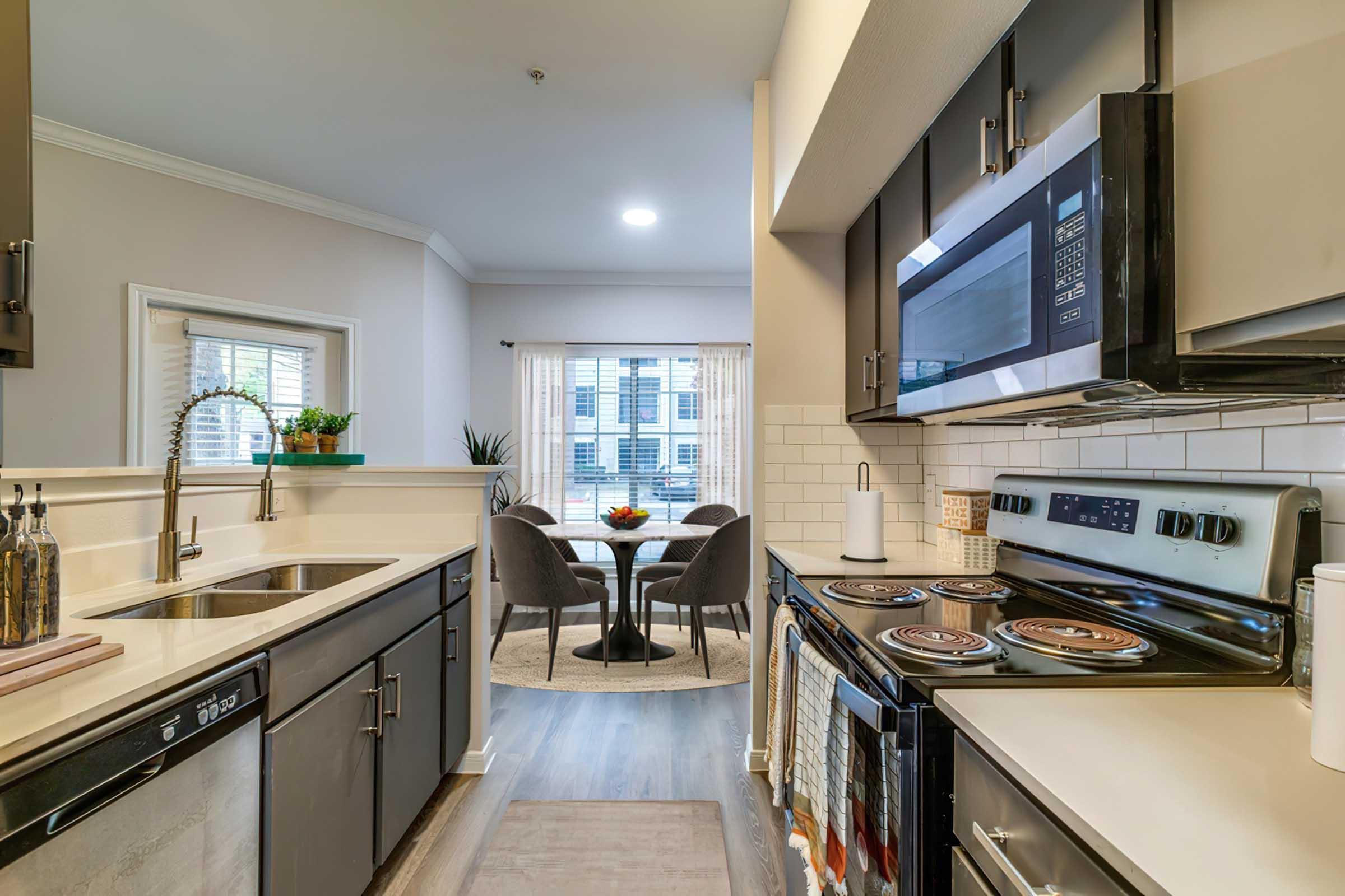 A modern kitchen featuring dark cabinets, a stainless steel stove and microwave, and a double sink. In the background, there's a dining area with a round table and chairs, illuminated by natural light from a window with sheer curtains. A bowl of fruit is placed on the table, enhancing the inviting atmosphere.