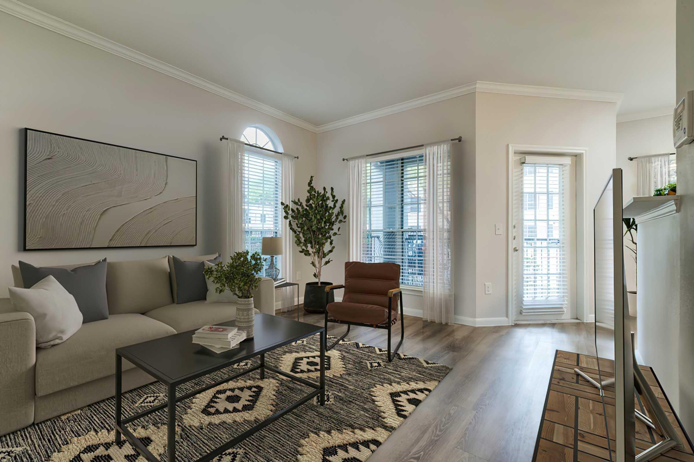 A cozy living room featuring a beige sofa with pillows, a dark coffee table, and an armchair. There is a large artwork on the wall, potted plants, and natural light coming through large windows with sheer curtains. A door leads to an outdoor area, and the floor has a patterned area rug.