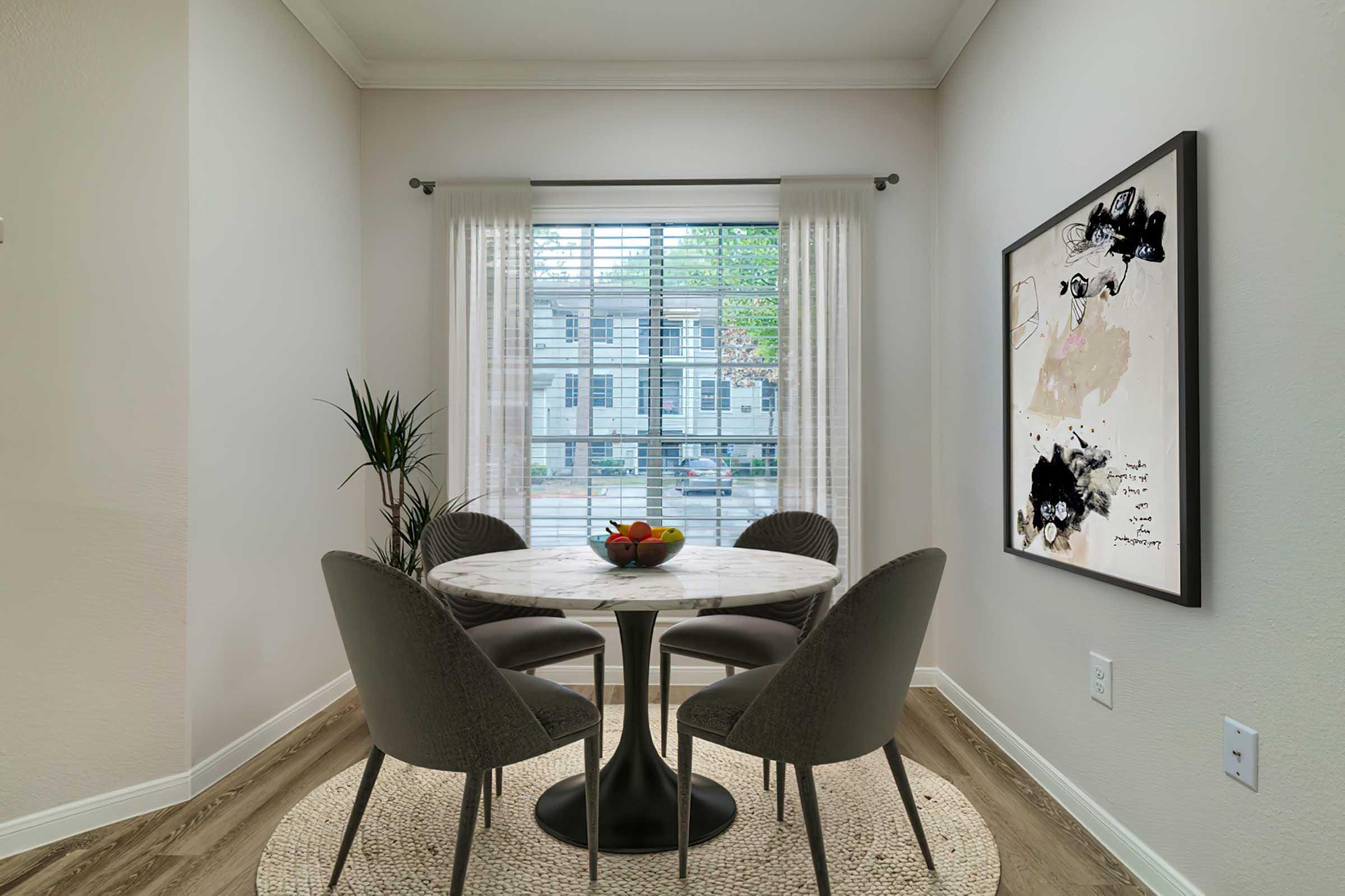 A cozy dining nook featuring a round marble table surrounded by four gray chairs. A small bowl of fruit sits on the table. The space is illuminated by natural light streaming through a window with blinds, and there’s a decorative framed artwork on the wall alongside a potted plant.