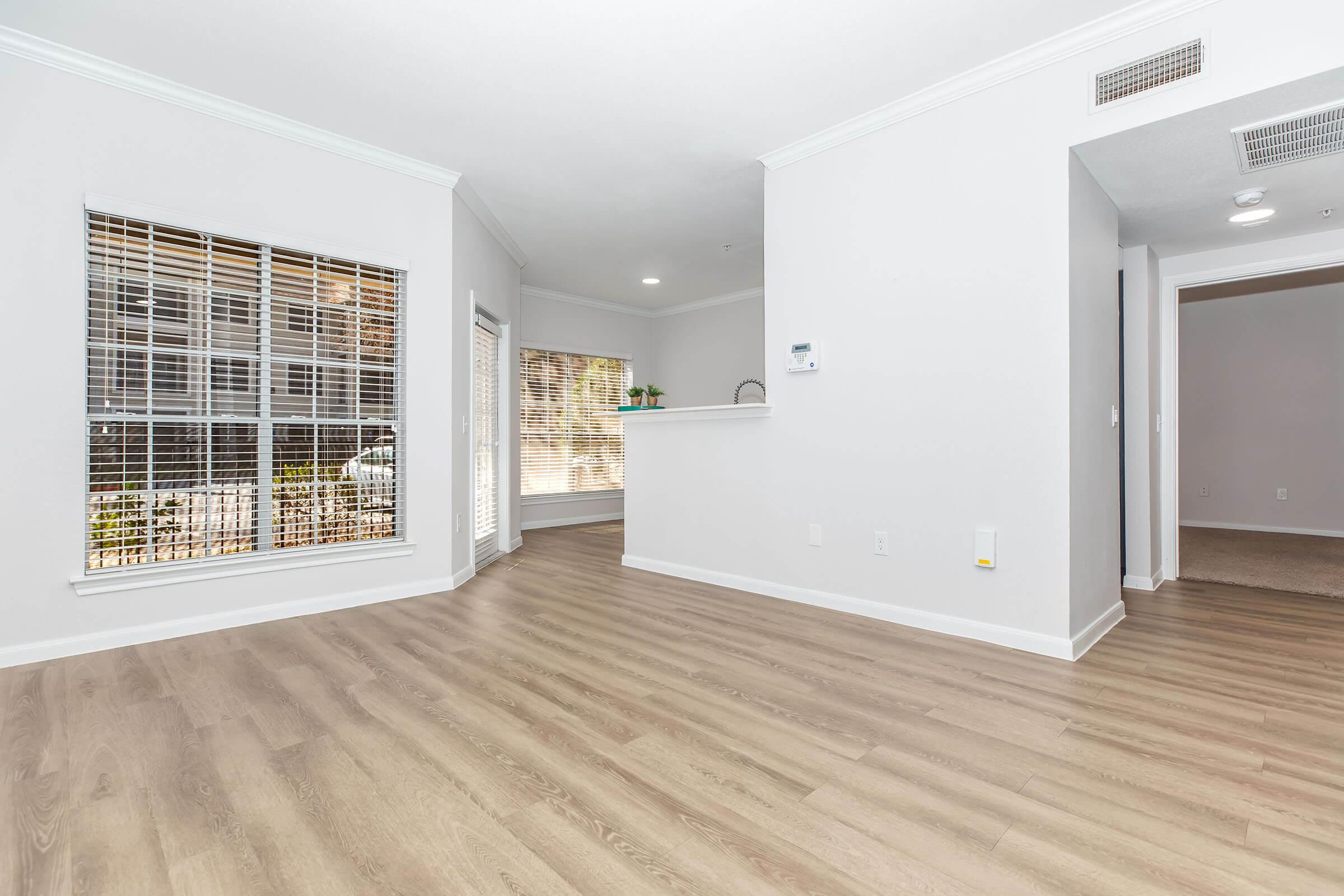 Spacious, modern living area with light-colored wood flooring, large windows with blinds, and a small kitchenette in the background. The walls are painted in a soft, neutral tone, creating a bright and inviting atmosphere. A doorway leads to another room, enhancing the open layout of the space.