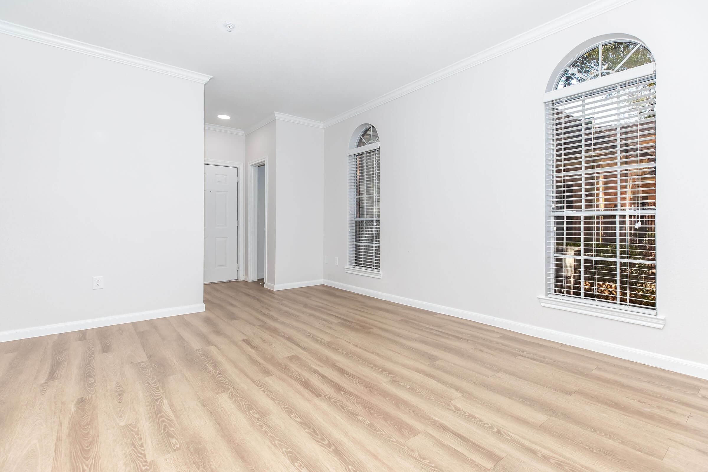 A spacious, empty room with light-colored wooden flooring and white walls. Two arched windows allow natural light to enter, featuring white blinds. There is a door on the left leading to another space, enhancing the open feel of the room. Ideal for various interior design possibilities.