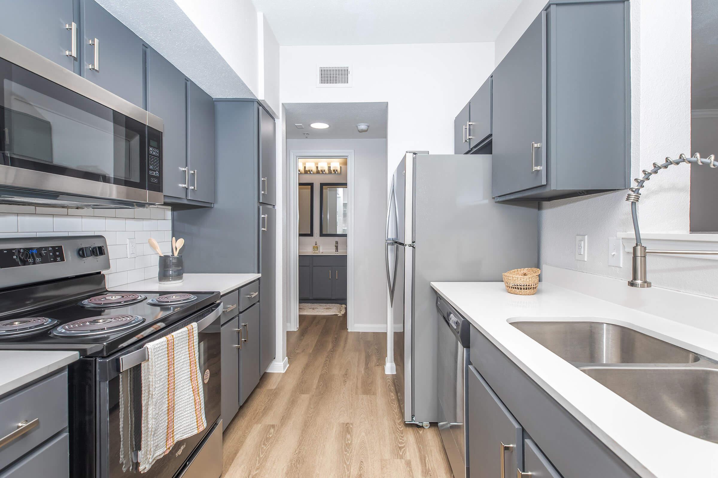 A modern kitchen featuring dark gray cabinetry, stainless steel appliances, and a white countertop. The kitchen includes a stove, microwave, and refrigerator. A hallway leads to a bathroom area, with natural light illuminating the space. The floor is finished in light wood laminate.