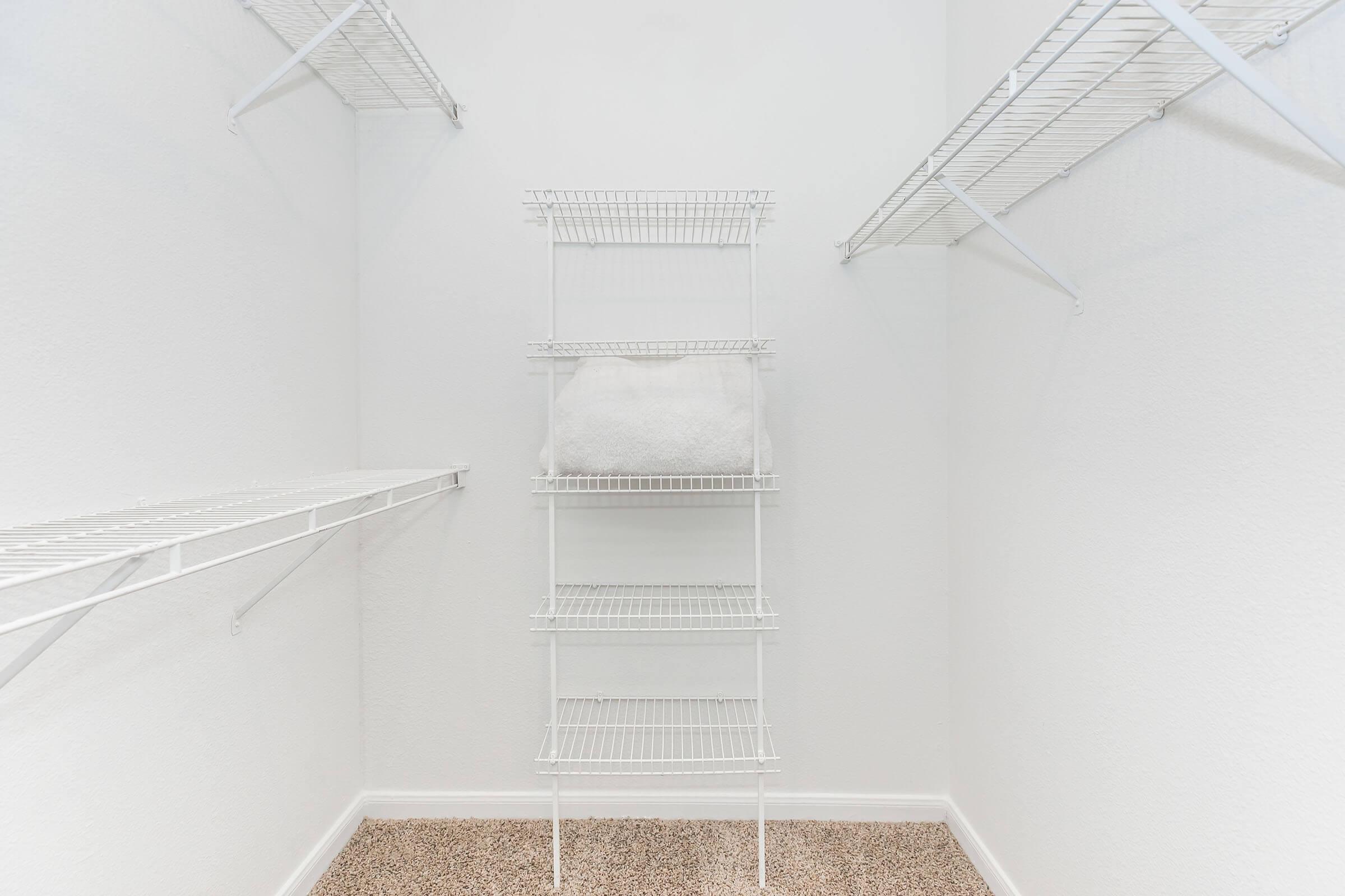 Empty closet with white wire shelving and a neatly folded white towel on a middle shelf. The walls are painted white, and the floor is covered in beige carpet. The overall space looks clean and organized, with ample storage options.