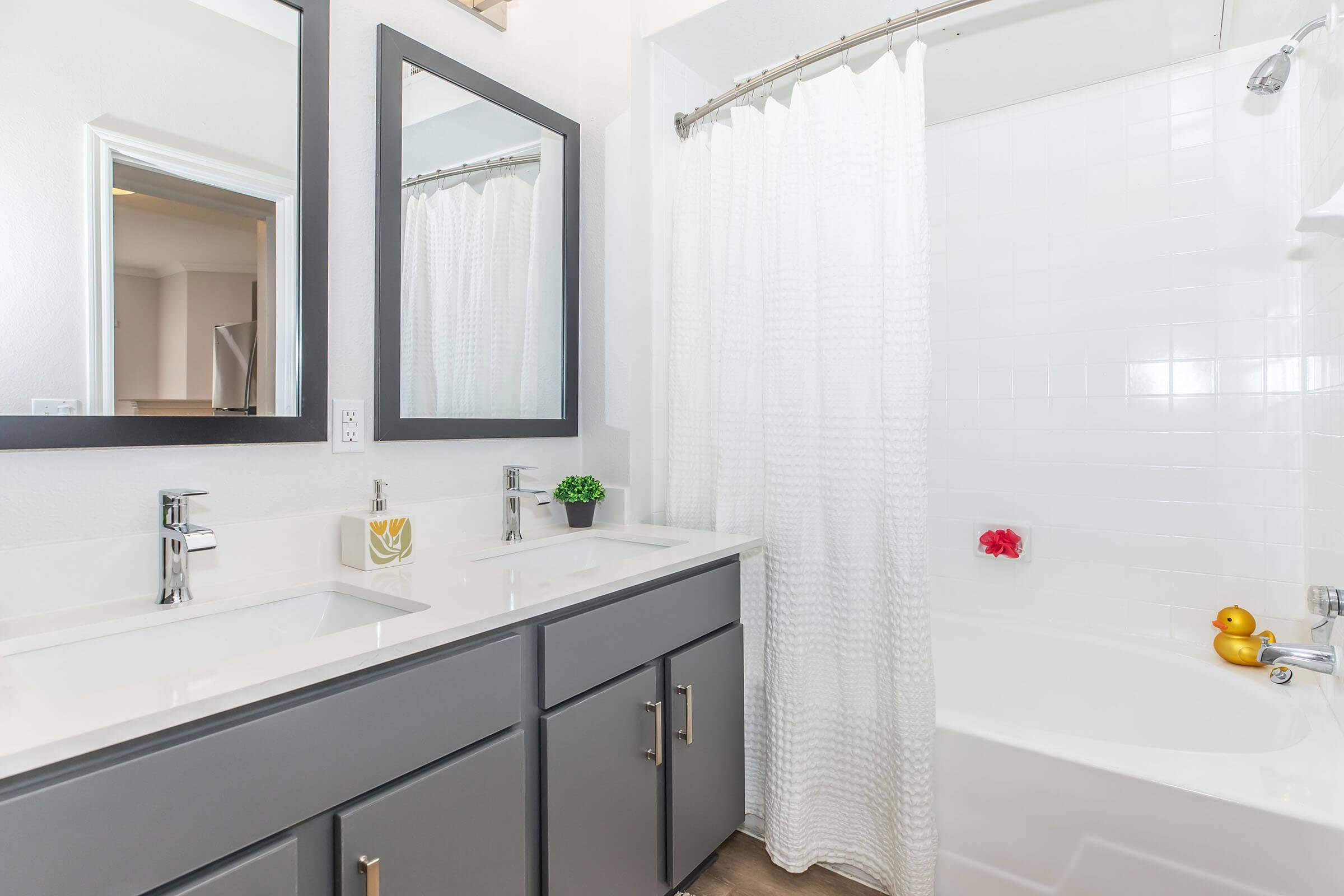A modern bathroom featuring a double vanity with dark gray cabinets, two mirrors, and stylish faucets. The space has white tiled walls and a white bathtub with a shower curtain. A small potted plant and decorative items, including a red flower and rubber duckies, add a touch of color and warmth.