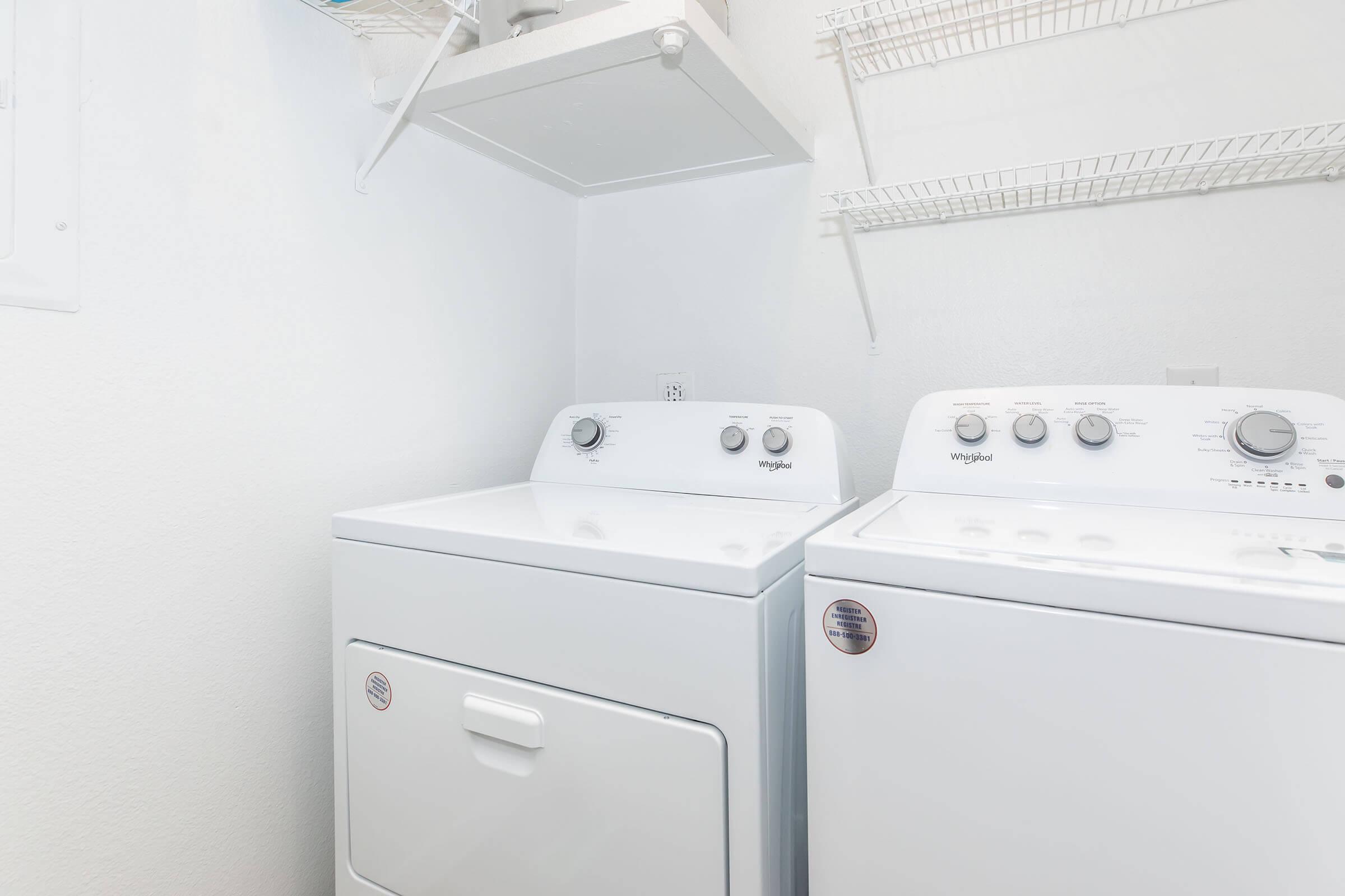 A clean, modern laundry room featuring a white washing machine and dryer side by side, with dials and buttons visible on the front. The walls are a light color, and there is a wire shelf above the appliances for additional storage.