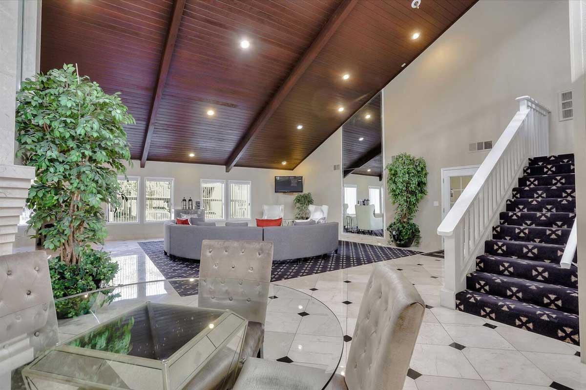 Interior of a spacious living room featuring high wooden beam ceilings, large windows, and stylish furniture. A cozy seating area with a circular gray sofa is complemented by decorative plants. A staircase with a white railing leads to an upper level, and the flooring is a mix of polished tiles and carpet.