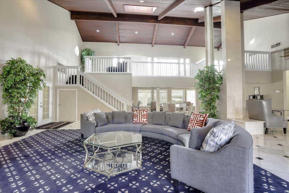 A spacious and modern lobby with high ceilings, featuring a curved gray sofa adorned with colorful pillows. A glass coffee table sits on a patterned rug, surrounded by lush indoor plants. In the background, there's a staircase leading to an upper level and additional seating areas visible.