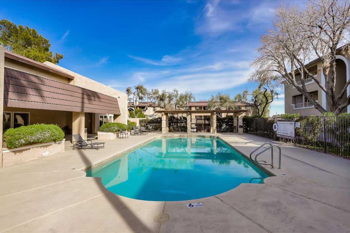 A clear swimming pool surrounded by lounge chairs and shaded areas, with trees and buildings in the background. The sky is bright blue with a few clouds, creating a serene outdoor atmosphere ideal for relaxation.