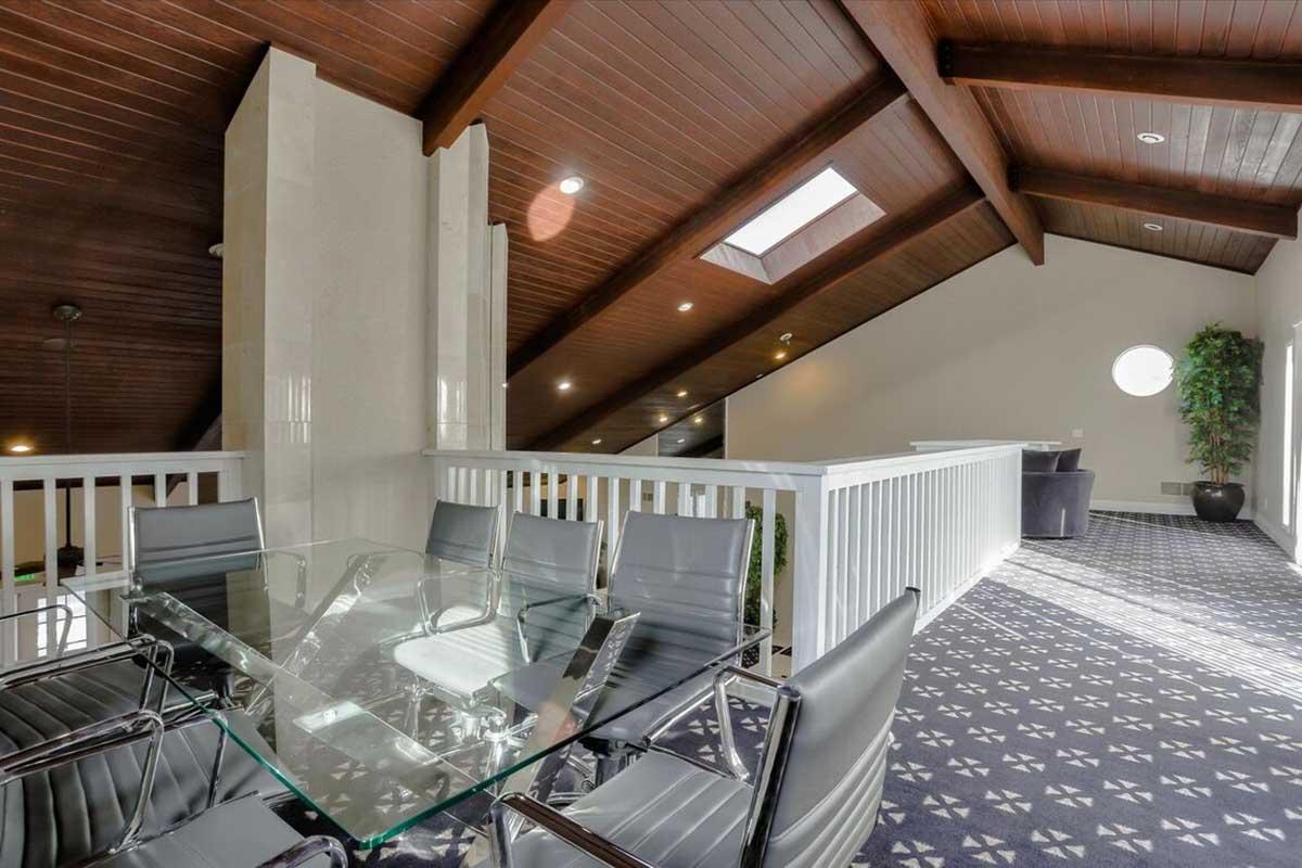 A modern interior space featuring a glass dining table and sleek chairs, set in a loft area with wooden beams and a high ceiling. Natural light enters through skylights and circular windows, complemented by indoor plants and a patterned carpet.