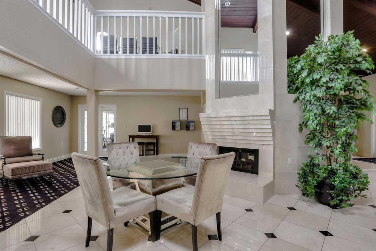 Modern living space featuring a glass dining table with upholstered chairs, elegant tile flooring, and a fireplace. The room is brightly lit with natural light from large windows, and a staircase leads to an upper level. Lush greenery adds a touch of nature to the contemporary decor.