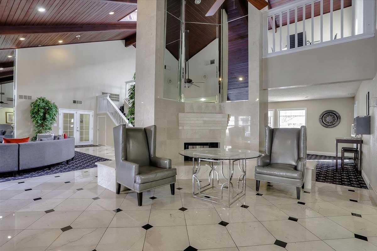 Modern interior of a spacious lobby with polished marble flooring. Two elegant gray chairs are placed beside a glass table, while large windows allow natural light to fill the space. A stylish staircase and greenery add to the atmosphere. The area features high ceilings and contemporary decor.
