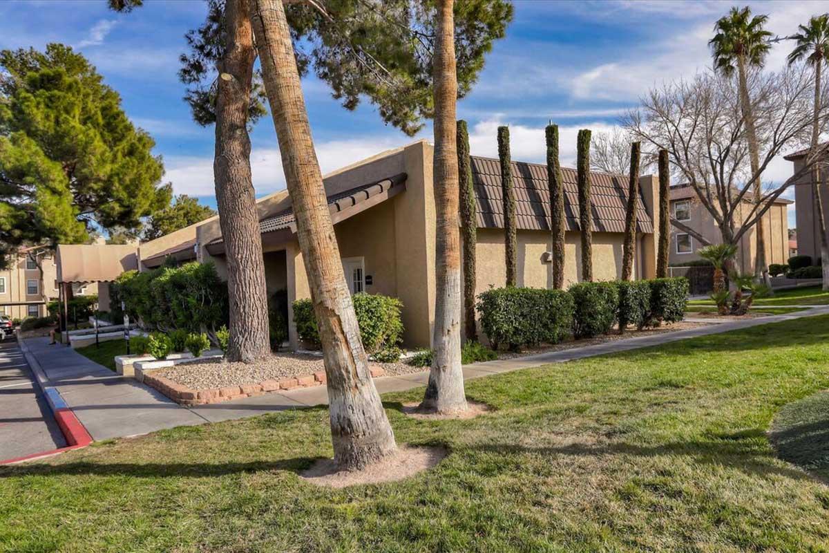 A landscaped area featuring a low building surrounded by neat shrubs and trees. The building has a sloped roof and is situated in a residential setting with a clear blue sky overhead. The greenery includes tall palm trees and well-maintained grass, contributing to a welcoming atmosphere.