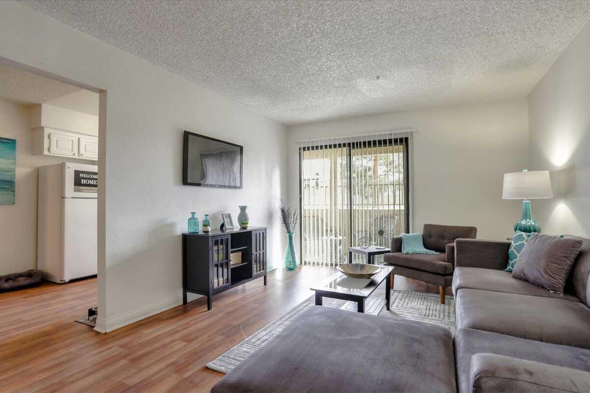 A cozy living room featuring a gray sectional sofa, a wooden coffee table, and a decorative cabinet. A large window with blinds lets in natural light, and decorative items in turquoise add color. In the background, a kitchen is visible with a refrigerator and a welcoming sign. The space has a warm, inviting atmosphere.