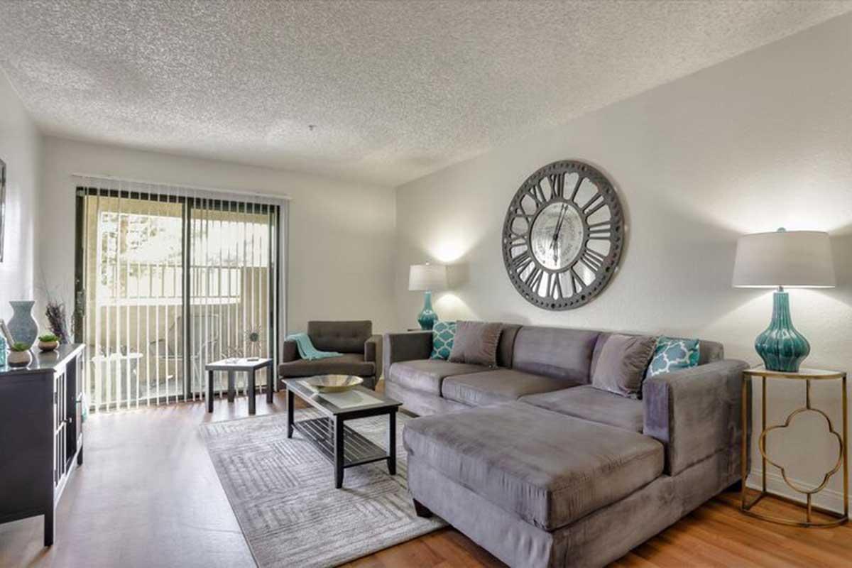 Cozy living room featuring a gray sectional sofa, decorative pillows, two armchairs, a glass coffee table, and a stylish wall clock. Large window with sliding doors allows natural light, complemented by neutral-toned walls and wooden flooring. Stylish decor elements enhance the inviting atmosphere.