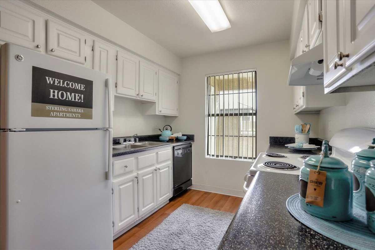 Bright kitchen featuring white cabinetry, stainless steel appliances, and a cozy decorative rug. A sign on the fridge reads "WELCOME HOME!" A window allows natural light, complemented by teal kitchen accessories and a stovetop with an overhead light.