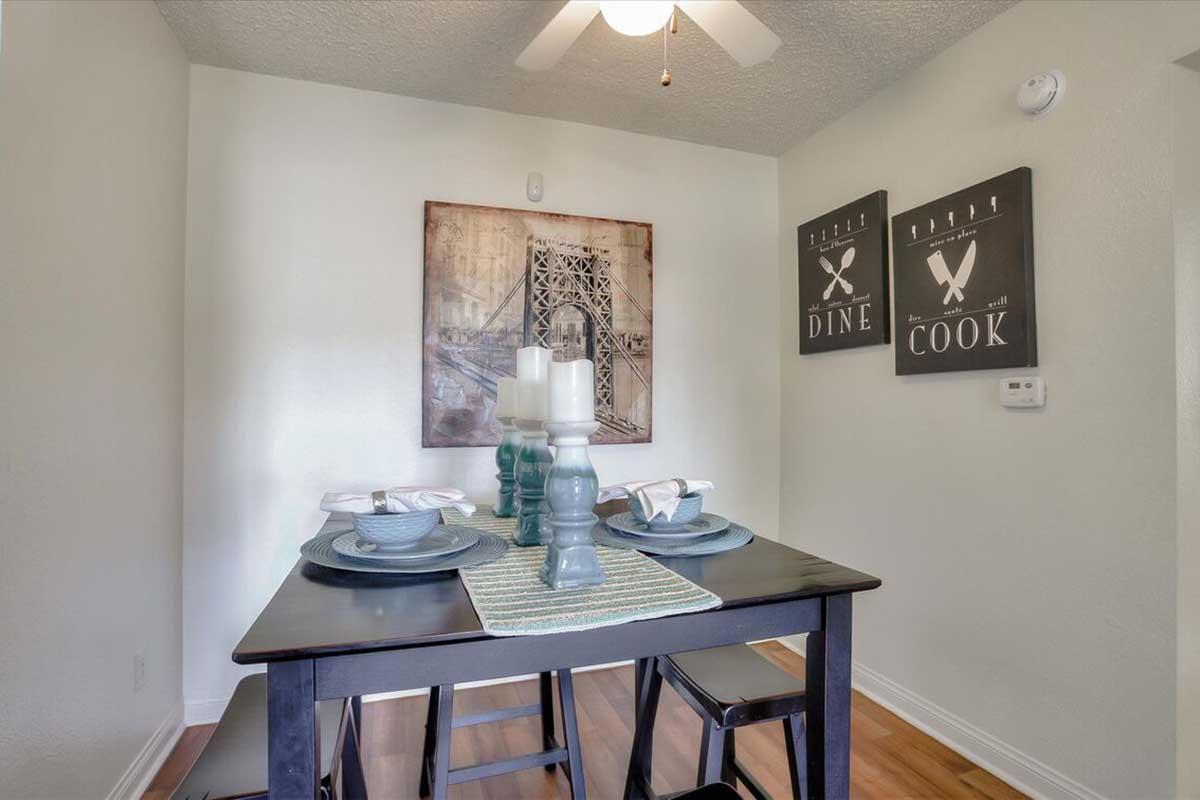 A dining area featuring a black table set for meals with blue plates and cutlery. A decorative runner is laid down the center, and there are tall candle holders in the middle. The walls are adorned with framed art and two signs that read "DINE" and "COOK." A ceiling fan is visible above.