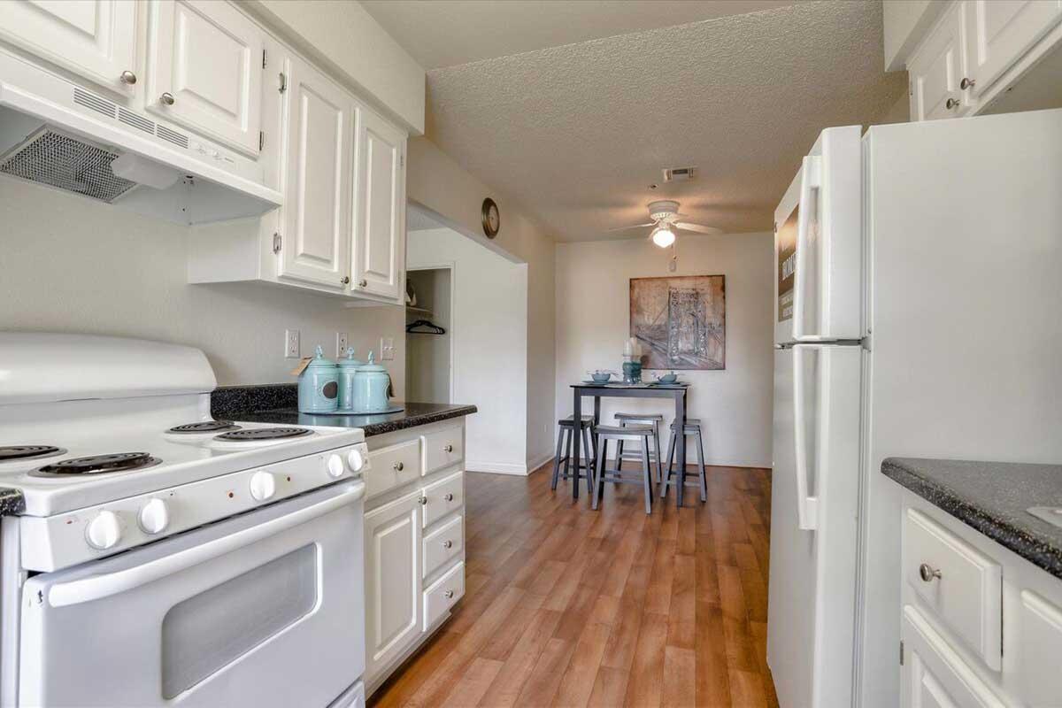 A bright and modern kitchen featuring white cabinets, a black countertop, and a stove. The kitchen opens up to a dining area with a small table and two chairs. Natural light enhances the space, highlighting the wooden floor and a wall-mounted artwork in the dining section.
