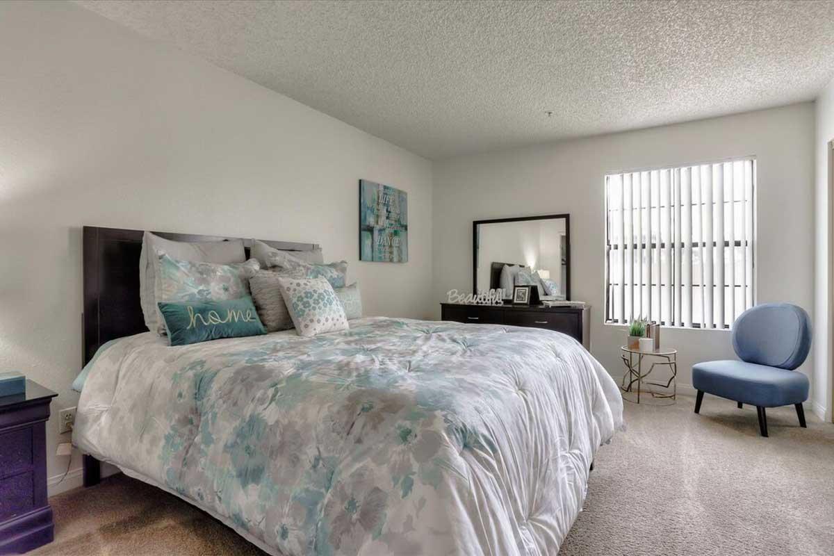 A cozy bedroom featuring a queen-sized bed with a floral-patterned comforter, decorative pillows, and a bedside table. A mirror and dresser are positioned against the wall, while a small blue accent chair is near a window with vertical blinds, providing natural light. The soft carpet adds warmth to the space.
