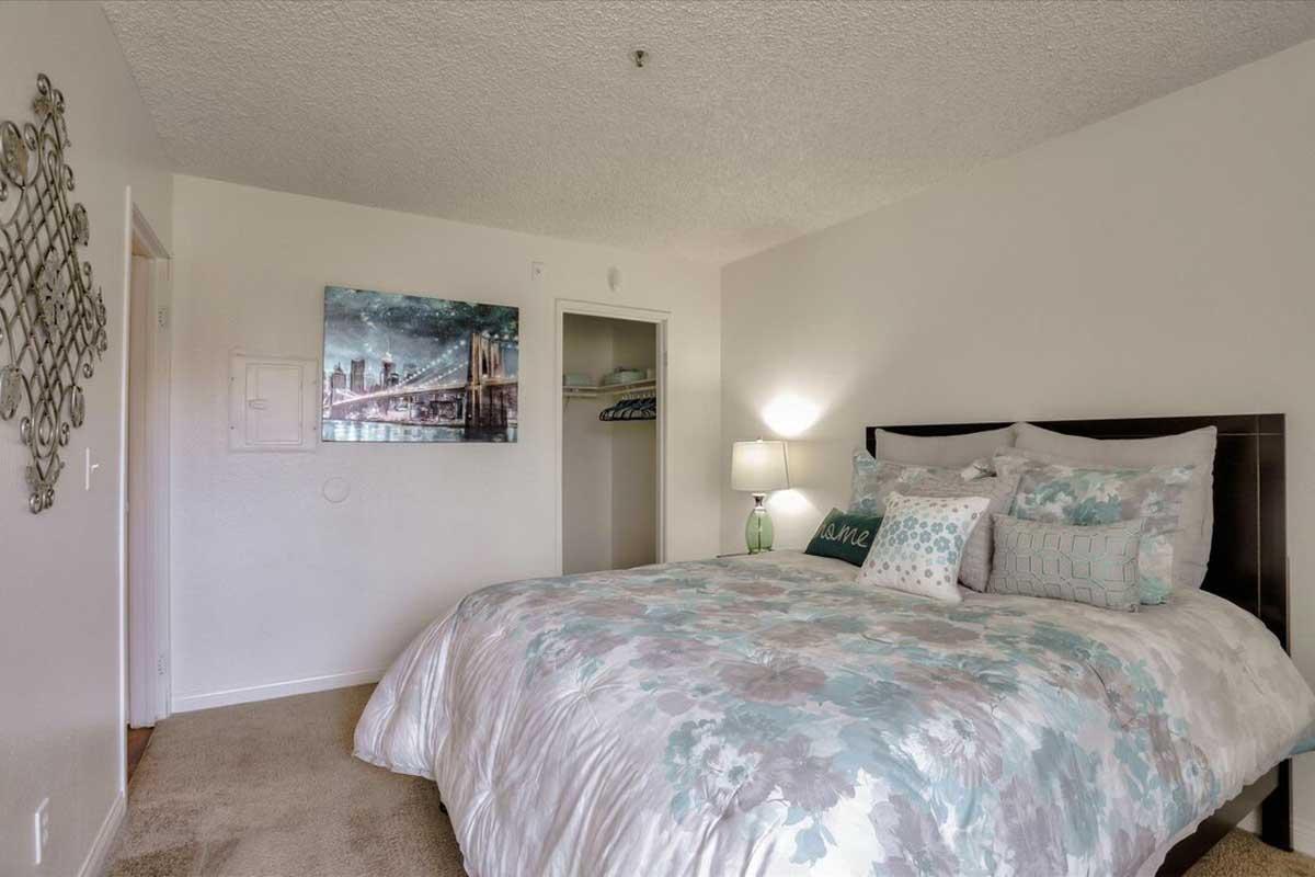 A cozy bedroom featuring a large bed with a floral-patterned comforter, decorative pillows, and a bedside lamp. A closet is visible on the right, and a wall art piece depicting a city skyline hangs above the bed. The room has a soft, neutral color scheme and carpeted flooring.