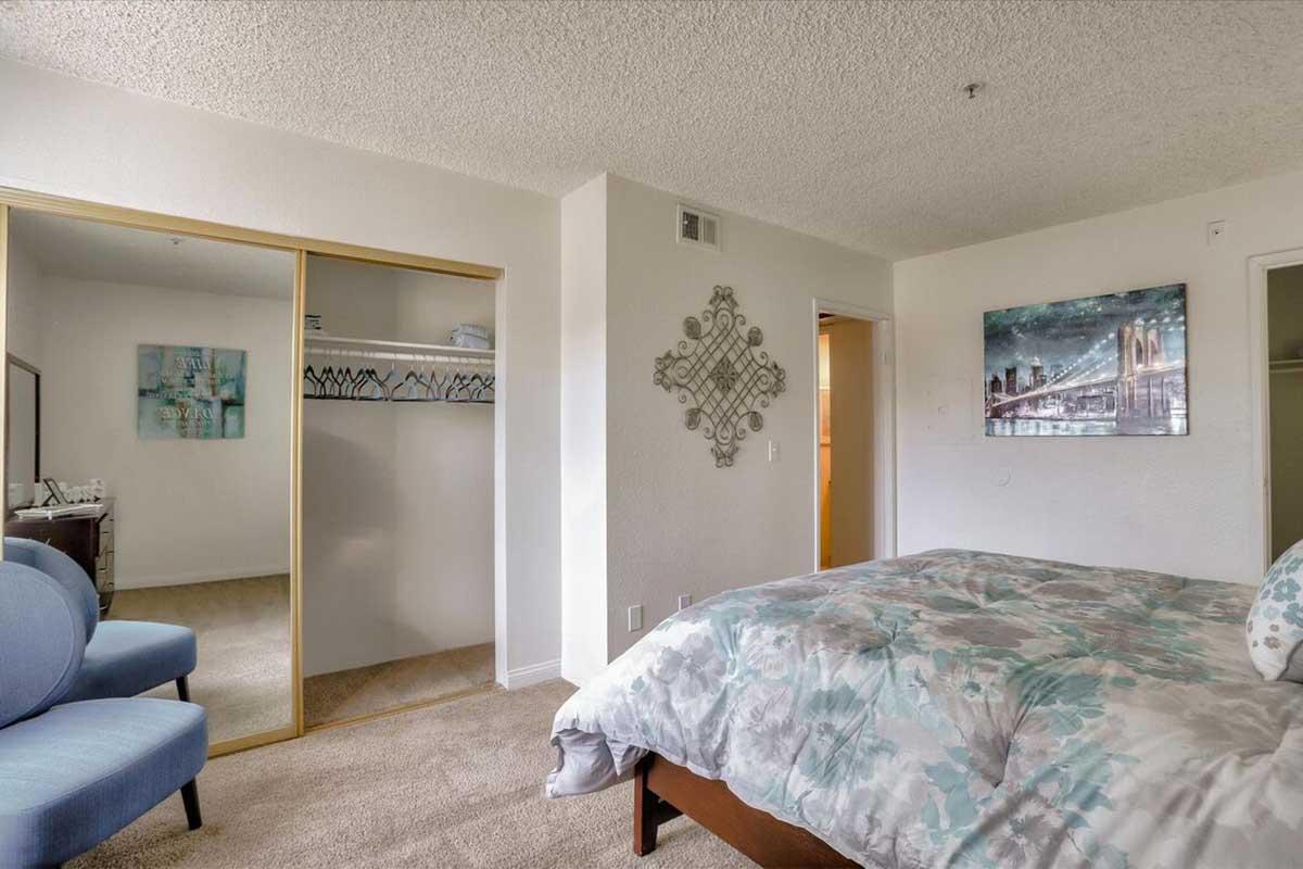 A cozy bedroom featuring a neatly made bed with a floral comforter, two blue accent chairs, and a decorative wall piece. A closet with sliding mirrors is visible, along with a light-colored wall and a painting of a bridge. The space is well-lit and has a comfortable, inviting atmosphere.