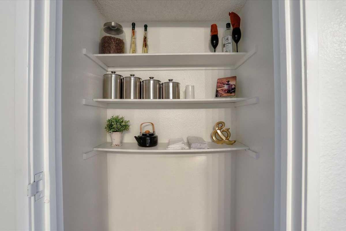 A neatly organized closet featuring multiple shelves. The upper shelves contain bottles, a small potted plant, and a decorative piece. The middle shelf holds metal containers and a small black pot, while the lower shelf has neatly folded towels. The closet has a clean, bright appearance.