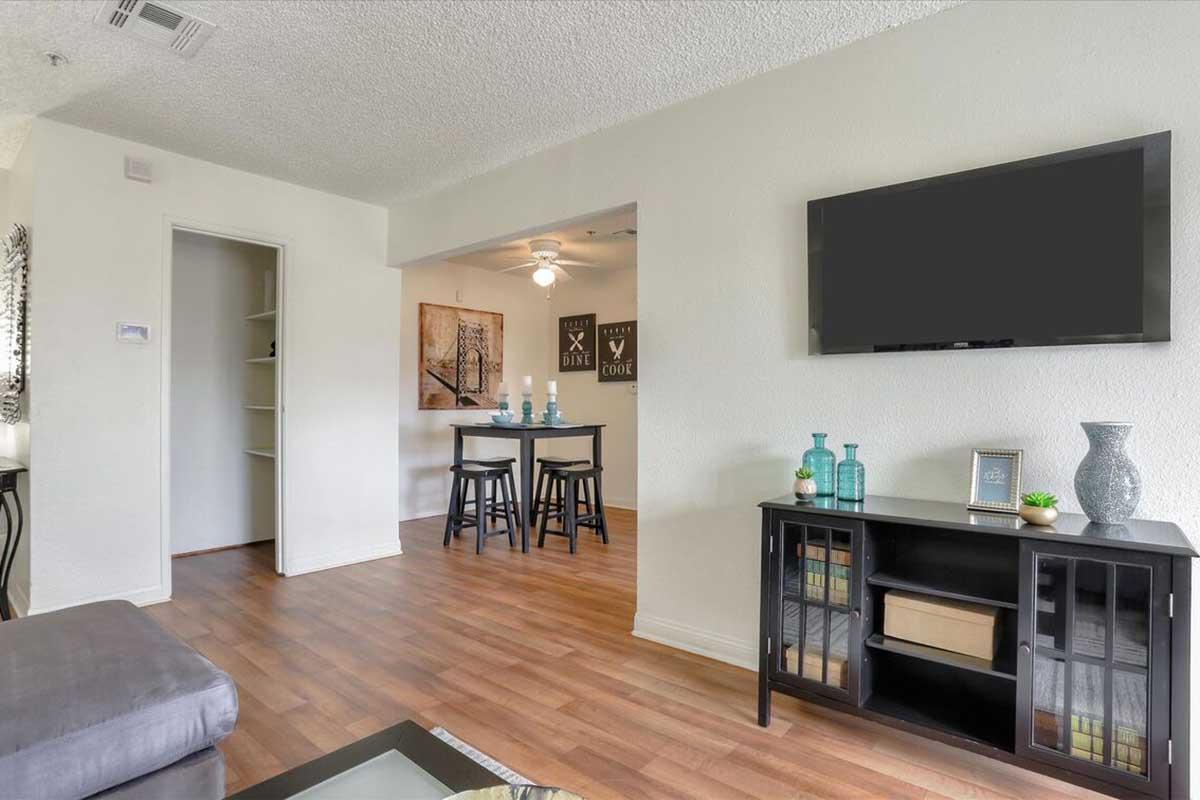 A cozy living space featuring a grey sofa, a coffee table, and a black cabinet with shelves. In the background, there's a dining area with a small table and chairs, complemented by decorative bottles. The room has wooden flooring and light-colored walls, creating a warm and inviting atmosphere.