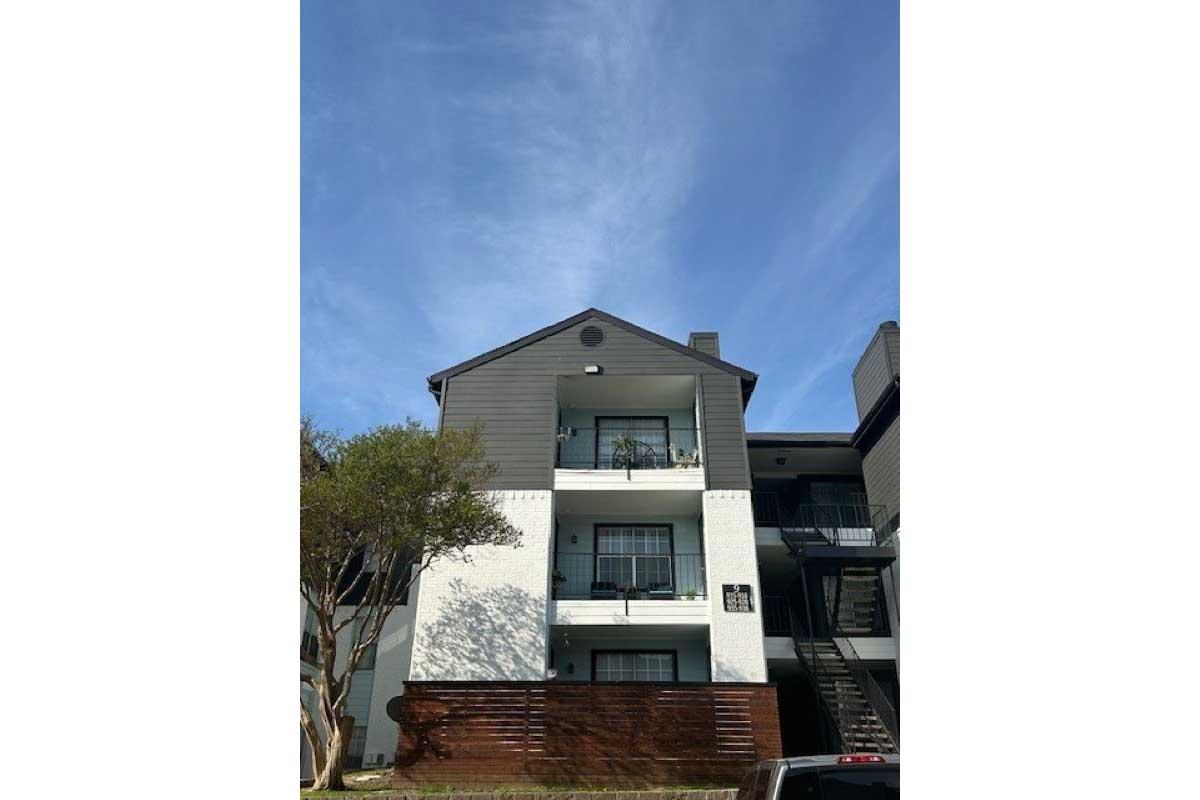 A three-story residential building featuring a combination of white and gray exteriors. The ground level has a wooden deck and visible stairs leading to the upper floors. The sky above is clear with light clouds.