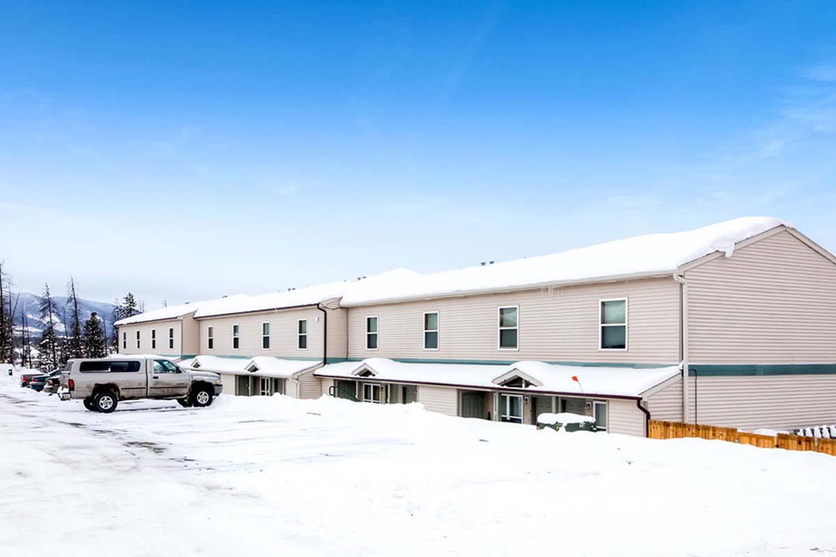 A row of beige multi-unit buildings covered in snow, with a parked white SUV in the foreground. The sky is clear and blue, and snow blankets the ground, suggesting winter conditions. The buildings have multiple windows and are flanked by trees in the background.