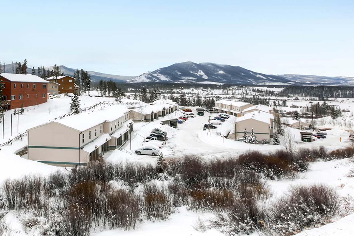 A snowy landscape featuring a row of buildings and parked cars in a winter setting. The scene is surrounded by hills and mountains in the background, with a clear sky and snow-covered ground, creating a tranquil, picturesque winter atmosphere.