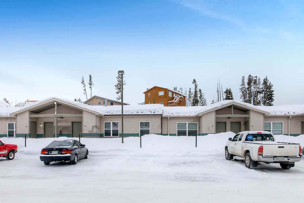 A row of single-story buildings covered in snow, with a clear blue sky above. Two parked cars, a red sedan and a white pickup truck, are in the foreground. Surrounding snow drifts and trees suggest a cold winter environment. The buildings feature sloped roofs and large windows.
