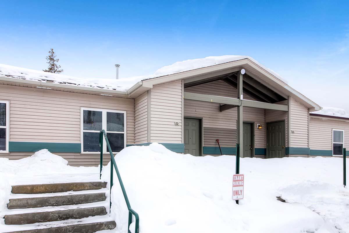 Single-story building with a sloped roof, surrounded by snow. It has green doors and windows on both sides, with a set of concrete steps leading to the entrance. A sign in front reads "ENTRY PARTIES ONLY," against a clear blue sky.