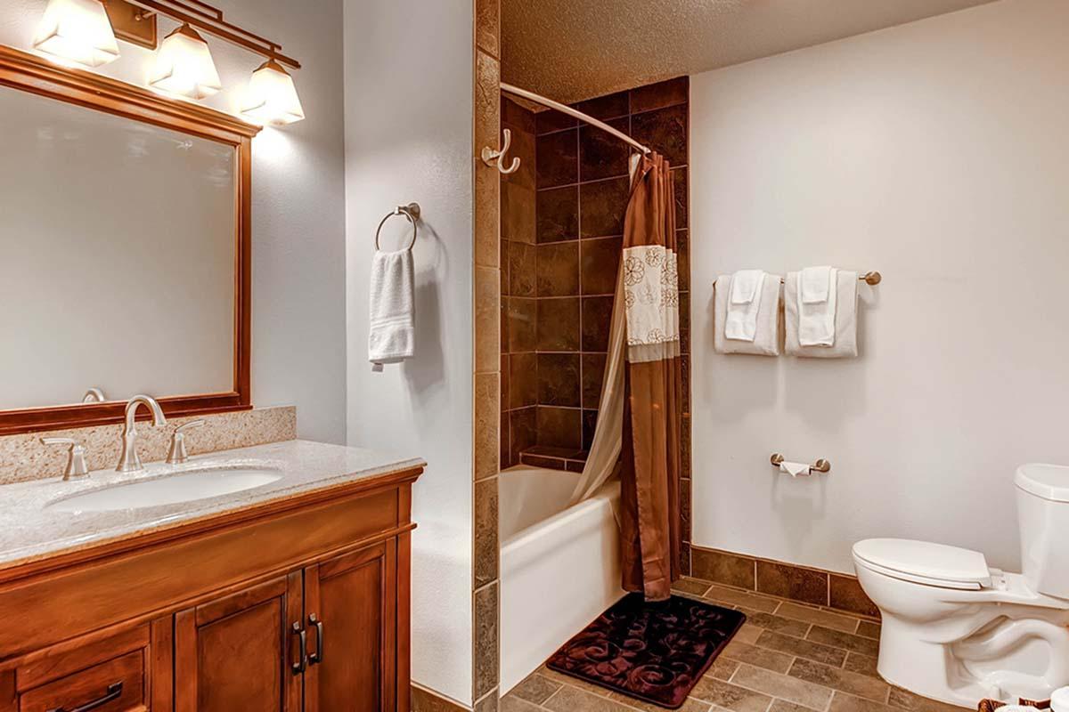 A modern bathroom featuring a wooden vanity with a sink, a bathtub with a shower curtain, a toilet, and neatly arranged towels on wall-mounted racks. Soft lighting from wall sconces illuminates the space. The flooring is tiled, complementing the overall decor.