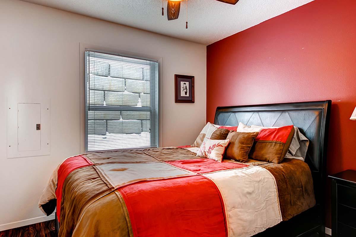 Cozy bedroom featuring a queen-sized bed with a colorful comforter in shades of red, brown, and beige. A window with blinds allows natural light, and a framed artwork hangs on the wall. The decor is warm and inviting, with wooden flooring enhancing the ambiance.