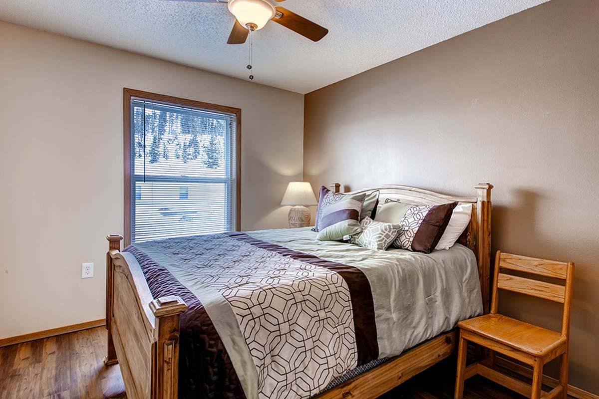 Cozy bedroom featuring a wooden bed with a patterned comforter, a bedside lamp, and a chair. A window allows natural light to fill the space, complementing the warm, neutral wall colors and wooden flooring.
