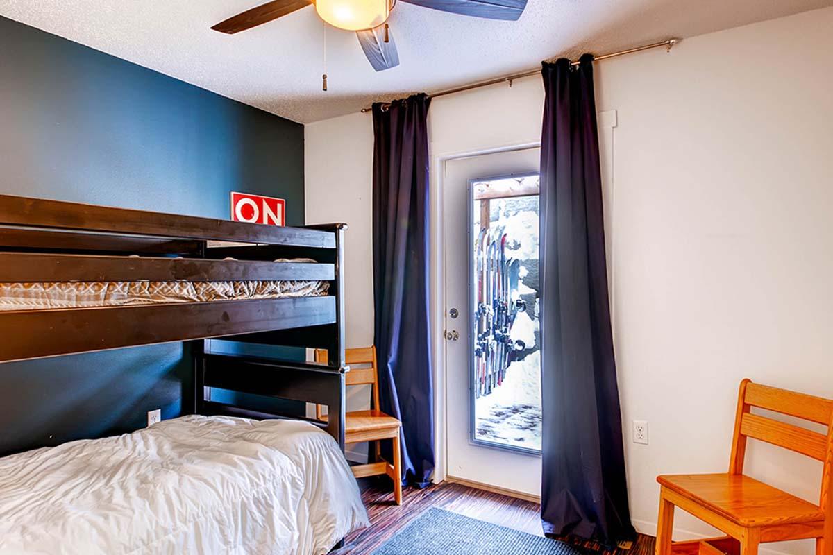 A cozy bedroom featuring a bunk bed with a white comforter, two wooden chairs, and a door leading to an outdoor area. The wall behind the bunk bed is painted dark blue, adding contrast to the neutral-colored walls. Curtains frame the door, allowing natural light to enter the room.