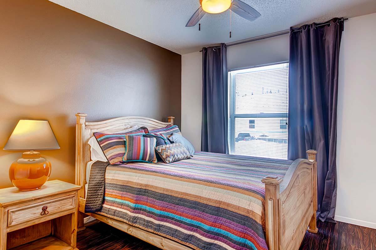 A cozy bedroom featuring a wooden bed with a colorful striped blanket, a nightstand with an orange lamp, and a window with light-filtering curtains. The walls are a warm brown, and the room has a calm, inviting atmosphere.