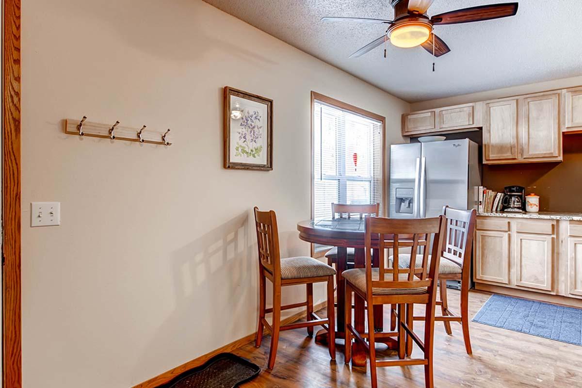 A cozy kitchen dining area featuring a small round table with four wooden chairs, a fan on the ceiling, a window providing natural light, and a glimpse of stainless steel appliances and cabinets in the background. A wall-mounted coat rack is visible on the left.