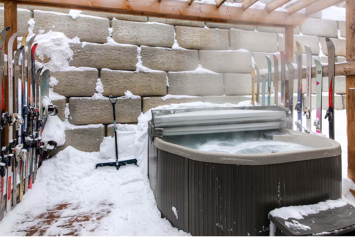 Hot tub surrounded by snow, with skis leaning against a stone wall. The scene features a cozy outdoor setting, perfect for relaxation after skiing, with steam rising from the hot water. Snow is piled nearby, and a shoveling tool rests beside the hot tub.