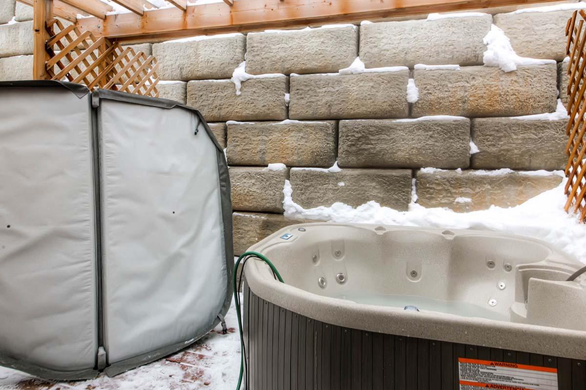 A snow-covered outdoor area featuring a hot tub and a gray enclosure for storage or privacy. The surrounding walls are made of large stone blocks, and the scene is set under a wooden pergola, providing some shelter from the elements.