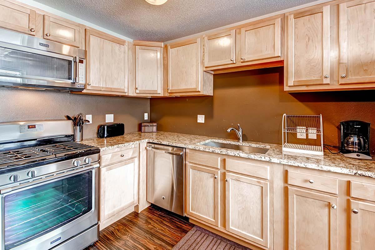 Modern kitchen with light wood cabinetry, granite countertops, stainless steel appliances including an oven and dishwasher, and a sink. The walls are painted dark brown, and there are small appliances such as a toaster and coffee maker on the countertop. The flooring is dark wood laminate.