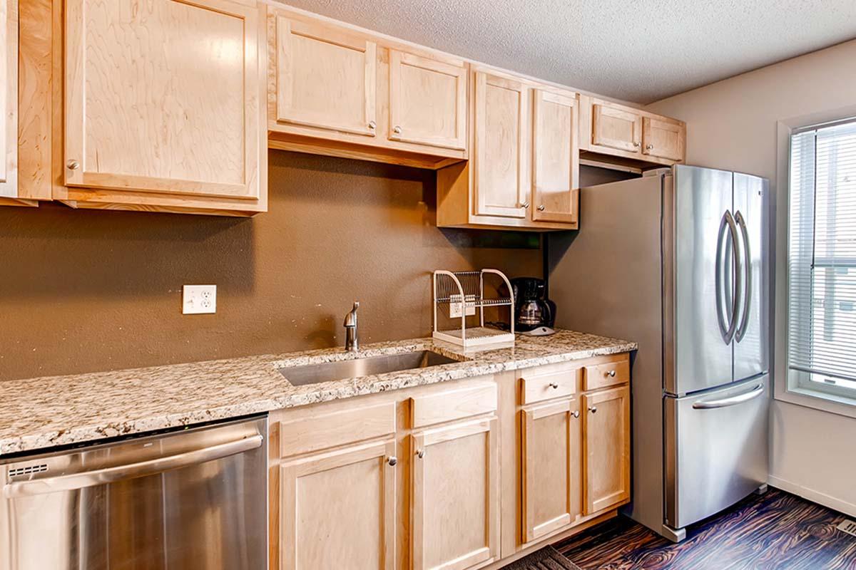 A modern kitchen featuring light wood cabinets, a stainless steel refrigerator, and a granite countertop. There is a double sink with a faucet, and the space is well-lit by natural light coming from a nearby window. A dishwasher is also visible under the counter.