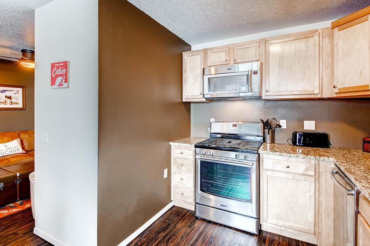 Modern kitchen featuring wooden cabinetry, a stainless steel stove, microwave, and a black toaster on the counter. A granite countertop adds elegance, and a cozy seating area is visible in the background. The wall has a decorative sign, enhancing the welcoming atmosphere.