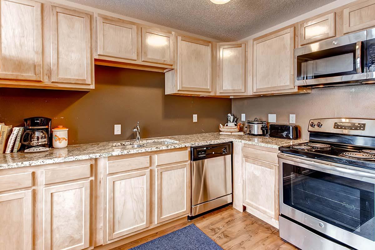 Modern kitchen featuring light wooden cabinets, a stainless steel dishwasher, and an oven with an electric stovetop. The countertop is covered with granite, and appliances like a microwave and coffee maker are visible. The space is well-lit and organized, with a rug on the floor.