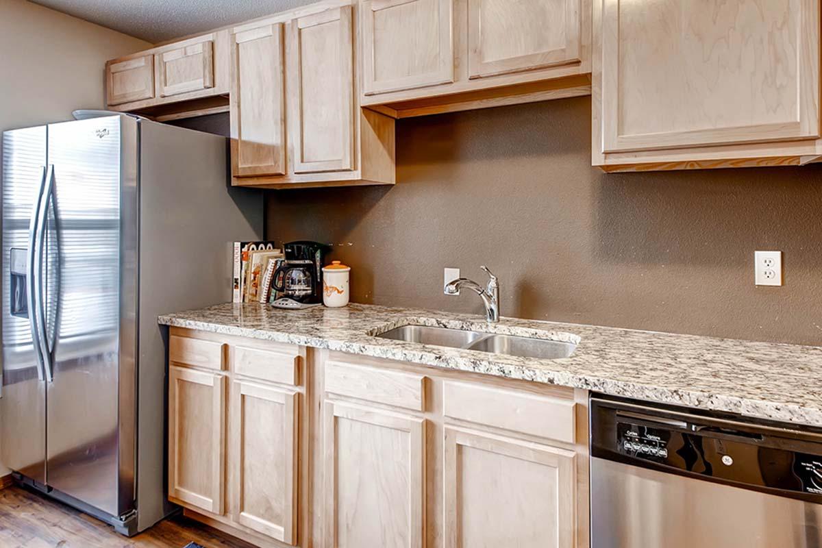 A modern kitchen featuring light wood cabinets, a stainless steel refrigerator, a silver dishwasher, and a granite countertop. The sink is positioned under a window, with various kitchen appliances and utensils neatly arranged on the countertop. The walls are painted a warm, neutral color.