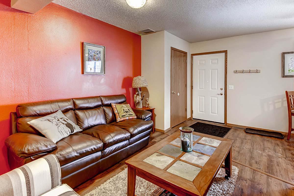 Living room with a brown leather sofa adorned with decorative pillows, a wooden coffee table with a stone inlay, and a lamp. The wall behind the sofa is painted red, while the adjacent wall is neutral. A closed door and a wall-mounted coat rack are visible, along with wood flooring and a cozy area rug.